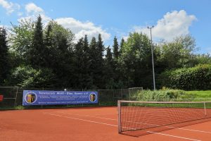 12 Meter breites Mesh-Banner des Tennisclub Weiß-Blau Hemer e.V. am Zaun der Tennisanlage. Das blaue Banner zeigt das vektorisierte Vereinslogo, den Schriftzug „Tennisclub Weiß-Blau Hemer e.V.“ sowie Informationen zu Flutlichttennis und Ganzjahresplätzen. Im Hintergrund sind die rote Sandplatz-Oberfläche, das Netz, weiße Bänke und eine Baumreihe unter blauem Himmel zu sehen.