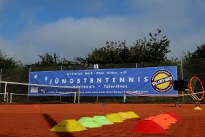 Großes blaues Mesh-Banner am Tenniszaun des TC Weiß-Blau Hemer e.V. mit der Aufschrift „Jüngstentennis – Knuddeltennis • Talentinos“. Das Banner wirbt für das Kindertennis-Programm, zeigt das offizielle gelb-rote Talentinos-Logo mit dem Slogan „Wir begeistern Kinder“.